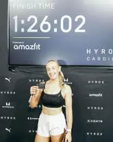 A photo of a participant at a Hyrox fitness event, standing in front of a branded backdrop displaying her finish time.