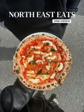 A top-down shot of a Margherita pizza held by a person outdoors, serving as a cover image for a 'North East Eats' monthly roundup. The image captures a casual, street-food vibe with a focus on the pizza itself.