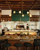 A professionally captured, wide-angle shot of a restaurant interior, showcasing an open kitchen with chefs at work and a neatly set dining table in the foreground. The warm, ambient lighting and symmetrical composition create an inviting and upscale atmosphere.