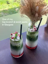 A bright, eye-level shot of two layered matcha drinks served in tall glasses, positioned on a table next to a window. The drinks feature distinct layers of red, white, and green, topped with crumbled meringue and herbs. A vase of dried flowers sits in the background, adding to the aesthetic.