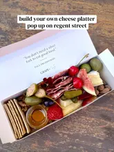 A high-quality, overhead shot of a 'build your own' cheese platter in a white box, featuring an assortment of salami, cheeses, fresh figs, cherries, strawberries, nuts, olives, pickles, and a small pot of honey, set against a rustic wooden surface.