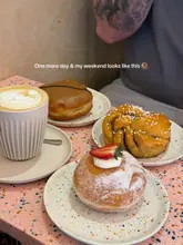 A bright, overhead flat-lay shot of a coffee and three pastries arranged on a speckled pink table, capturing a relaxed weekend cafe atmosphere.