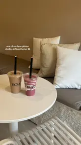 A clean, minimalist, and aesthetically pleasing shot of two smoothies placed on a white table in front of a neutral-toned seating area with cushions, captured inside a Pilates studio.