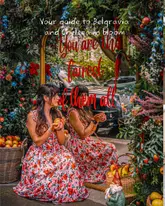 A high-quality, thematic photograph capturing a floral installation in London, likely part of the Chelsea or Belgravia in Bloom events. The image features a creator posing in front of a mirror surrounded by flowers and apples, creating a fairytale aesthetic that is visually engaging and on-brand for the event.