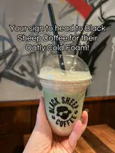 A close-up, eye-level shot of a hand holding a plastic cup of iced coffee with cold foam from Black Sheep Coffee, set against a graffiti-style wall.