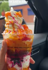 A short, energetic vlog-style video featuring a creator trying a 'Mango Fusion Sundae' and a box of fried chicken in a car, highlighting the arrival of a new summer dessert in Manchester.