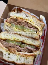 A high-quality, close-up shot of a layered sandwich served in a cardboard takeaway box. The lighting is natural and flattering, effectively highlighting the textures of the focaccia, meat, and pickles. The composition is tight and focused, making the food the clear subject, which is ideal for a casual street food or takeaway venue.