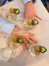 A top-down, overhead shot of four martini glasses arranged on a table, with hands visible in the frame. The image features a text overlay describing the drinks as the 'final boss of London's martini scene'.
