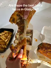 A close-up, first-person perspective shot of a birria taco being pulled apart to showcase a cheese stretch, set against a backdrop of takeaway containers and fries on a marble surface.