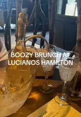 A vlog-style video showcasing a 'Fizz Club' brunch experience at Luciano's, featuring pasta dishes, garlic bread, chips, and non-alcoholic sparkling wine, accompanied by live saxophone music and a lively atmosphere.