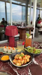 A high-quality, well-composed shot of a table spread at a rooftop restaurant, featuring sushi, edamame, fried shrimp, and cocktails. The image captures the atmosphere of a premium dining experience with a view.