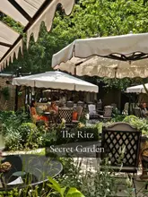A wide-angle shot of the outdoor garden terrace at The Ritz, featuring white umbrellas, lush greenery, and patrons seated at tables.