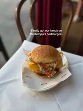 A high-quality, well-composed close-up shot of a tempura cod burger served on branded paper on a white tablecloth. The lighting is soft and natural, highlighting the textures of the bun and the fish, with a shallow depth of field that keeps the focus entirely on the food.