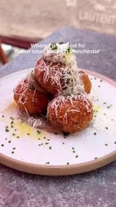 A short, music-led reel showcasing an Italian restaurant called Bruco in Manchester. The video features a quick shot of the creator enjoying a drink at an outdoor table, followed by close-ups of dishes like arancini, garlic butter flatbread, and fresh fish. It also includes a brief look at the menu, the restaurant interior, and the exterior.