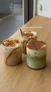 A high-quality, well-lit photograph of three iced drinks—two coffee-based and one matcha-based—arranged on a wooden table. The image uses natural window light to highlight the textures of the drinks, including the toppings and the gradient of the matcha.