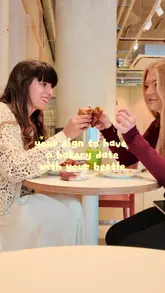 A candid, lifestyle-focused shot of two friends holding up a ham and cheese croissant in a cafe setting, featuring text overlay encouraging a bakery date.