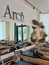 A bright, clean, and aesthetically pleasing shot of a Pilates studio featuring rows of reformer machines. The space is well-lit with natural light coming through the windows, and the decor is minimalist and modern, accented by a balloon arch.