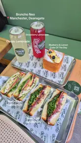 A clean, well-composed flat lay photograph showcasing a selection of sandwiches, a cube pastry, and canned drinks from Bruncho Neo in Manchester. The image effectively uses natural lighting and a simple, branded background to highlight the menu items, making it an appealing promotional shot for a casual lunch or brunch spot.