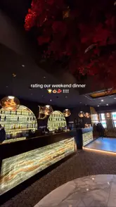 A wide-angle, atmospheric shot of a modern, upscale restaurant and bar interior, featuring warm accent lighting, illuminated shelving, and decorative red floral elements on the ceiling.
