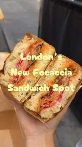 A high-quality, close-up shot of a freshly prepared focaccia sandwich held in hand, likely at a market stall. The image effectively showcases the texture of the bread and the vibrant fillings, making it highly appealing for a quick lunch option.